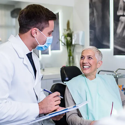 Doctor with clipboard in mask looking at smiling patient in dental chair.