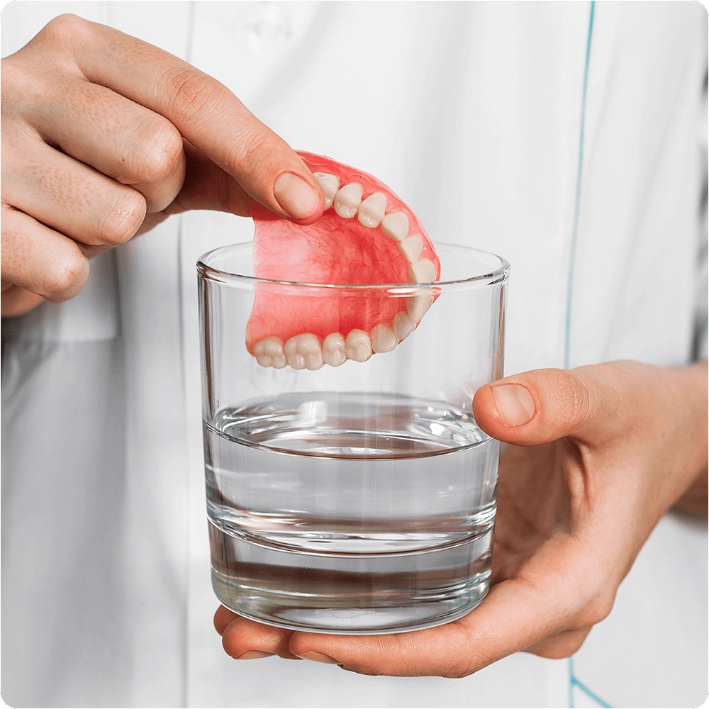 Hand grasping denture and placing it in a glass of water.